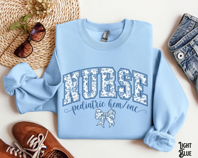May include: Light blue sweatshirt with the word "NURSE" in large, floral-patterned letters. Below, it reads "pediatric hem/onc" with a bow graphic. The sweatshirt is surrounded by accessories, including sunglasses and shoes.