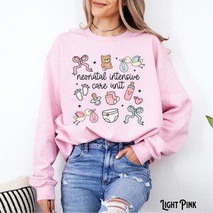 Op de afbeelding: Lichtroze sweatshirt met de tekst "neonatal intensive care unit" en baby-thema illustraties, waaronder een teddybeer, een speen, een babyfles, een luier en een ooievaar.