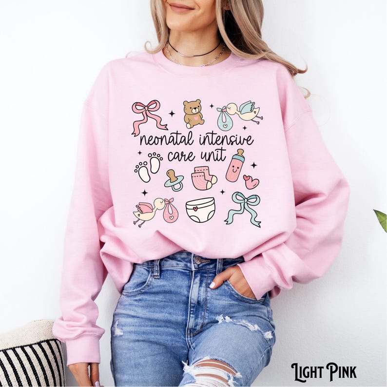 Op de afbeelding: Lichtroze sweatshirt met de tekst "neonatal intensive care unit" en baby-thema illustraties, waaronder een teddybeer, een speen, een babyfles, een luier en een ooievaar.