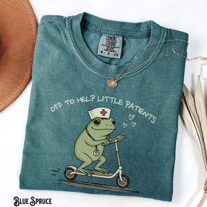 May include: Teal Comfort Colors t-shirt featuring a cartoon frog wearing a nurse's hat and riding a scooter. The text "OFF TO HELP LITTLE PATIENTS" is printed above the frog. A gold necklace is draped across the shirt.