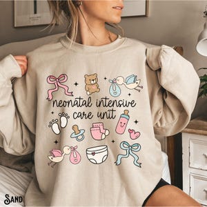Op de afbeelding: Een beige sweatshirt met een grafisch ontwerp van babygerelateerde iconen en de tekst "neonatal intensive care unit".