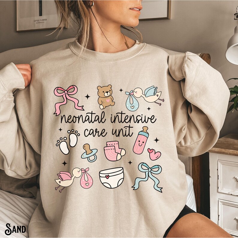 Op de afbeelding: Een beige sweatshirt met een grafisch ontwerp van babygerelateerde iconen en de tekst "neonatal intensive care unit".