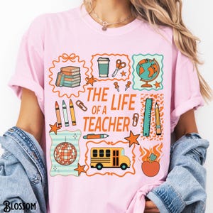 May include: Pink t-shirt with a colorful graphic design featuring school-related icons and the text "THE LIFE OF A TEACHER". The design includes a globe, school bus, apple, and other school supplies.