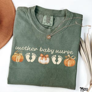 May include: A moss green t-shirt featuring the text "mother baby nurse" and pumpkin and footprint illustrations. A gold necklace with a pendant is draped across the shirt.