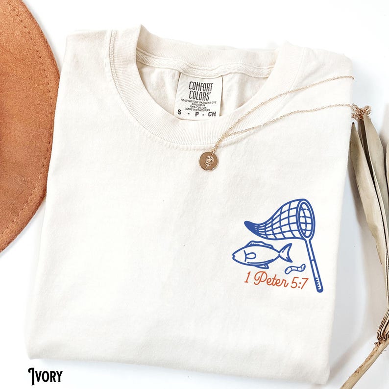 K&ouml;nnte beinhalten: Elfenbeinfarbenes T-Shirt mit blauem Fisch- und Netz-Motiv und dem Text "1 Peter 5:7" in Rot. Eine goldene Halskette mit Anh&auml;nger liegt &uuml;ber dem Shirt. Das Shirt ist mit "Comfort Colors" gekennzeichnet.