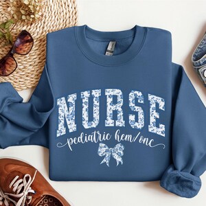May include: Indigo blue sweatshirt with the word "NURSE" in large white letters with a floral pattern. Below the word "NURSE" is the text "pediatric hem/onc" and a bow graphic. The sweatshirt is a crew neck style.