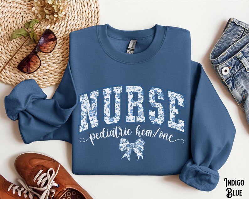 May include: Indigo blue sweatshirt with the word "NURSE" in large white letters with a floral pattern. Below the word "NURSE" is the text "pediatric hem/onc" and a bow graphic. The sweatshirt is a crew neck style.