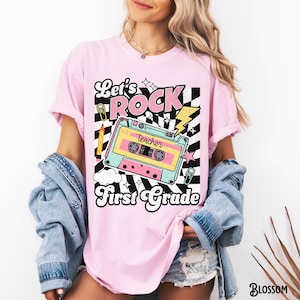 May include: Pink t-shirt with a retro design featuring a cassette tape, lightning bolt, and the words "Let's Rock First Grade." The design includes a pencil, paperclip, and stars against a black and white checkered background. The shirt is a light pink color.