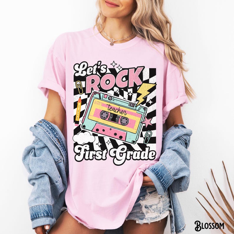 May include: Pink t-shirt with a retro design featuring a cassette tape, lightning bolt, and the words "Let's Rock First Grade." The design includes a pencil, paperclip, and stars against a black and white checkered background. The shirt is a light pink color.