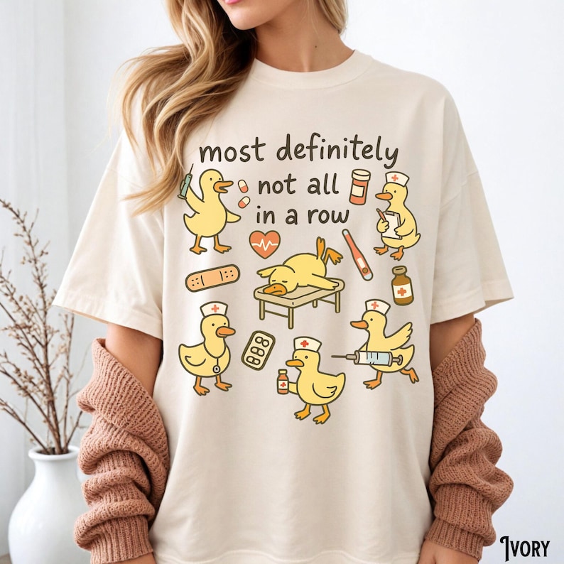 Puede incluir: Camiseta de color marfil con la frase "most definitely not all in a row" y dibujos de patos de dibujos animados en roles m&eacute;dicos. Los patos se representan con herramientas m&eacute;dicas como jeringas, vendas y estetoscopios. La camiseta es de estilo casual y unisex.