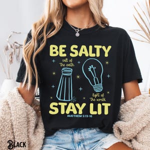 Puede incluir: Camiseta negra con el texto "BE SALTY STAY LIT" en amarillo, con un dise&ntilde;o de salero y bombilla. Tambi&eacute;n se incluye el texto "sal de la tierra" y "luz del mundo". El vers&iacute;culo b&iacute;blico Mateo 5:13-16 est&aacute; impreso debajo.