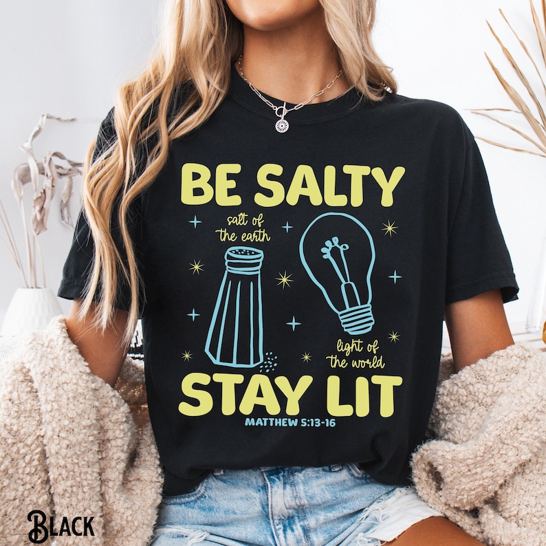 Puede incluir: Camiseta negra con el texto "BE SALTY STAY LIT" en amarillo, con un dise&ntilde;o de salero y bombilla. Tambi&eacute;n se incluye el texto "sal de la tierra" y "luz del mundo". El vers&iacute;culo b&iacute;blico Mateo 5:13-16 est&aacute; impreso debajo.