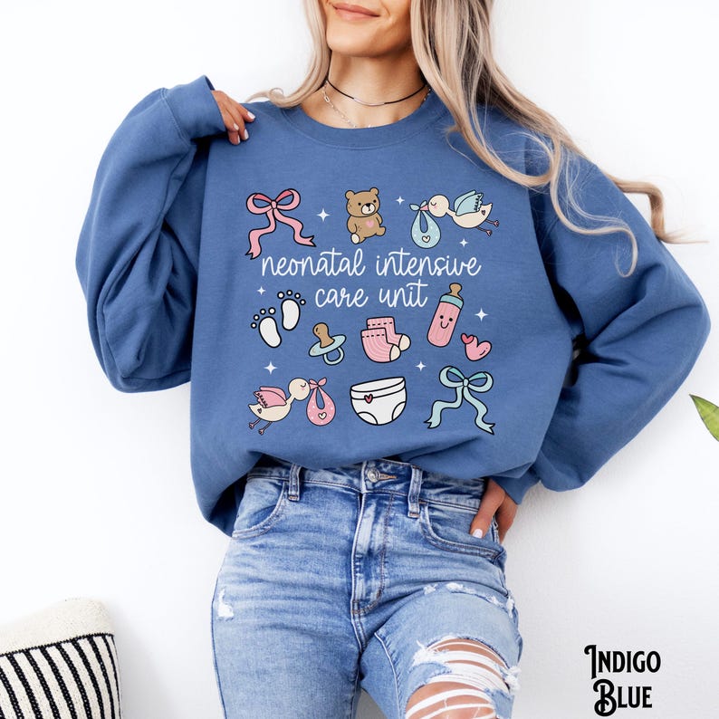 Op de afbeelding: Een blauw sweatshirt met de tekst "neonatal intensive care unit" en baby-thema illustraties, waaronder een teddybeer, een speen, een babyfles en een ooievaar.