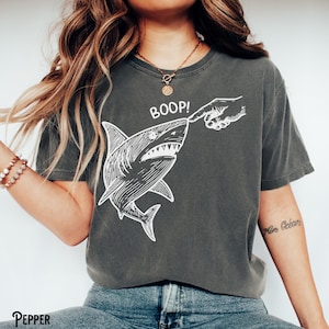 Pu&ograve; includere: T-shirt grigio scuro con un'illustrazione bianca di uno squalo che viene toccato sul naso, con la scritta "BOOP!" sopra. La maglietta ha una vestibilit&agrave; ampia.