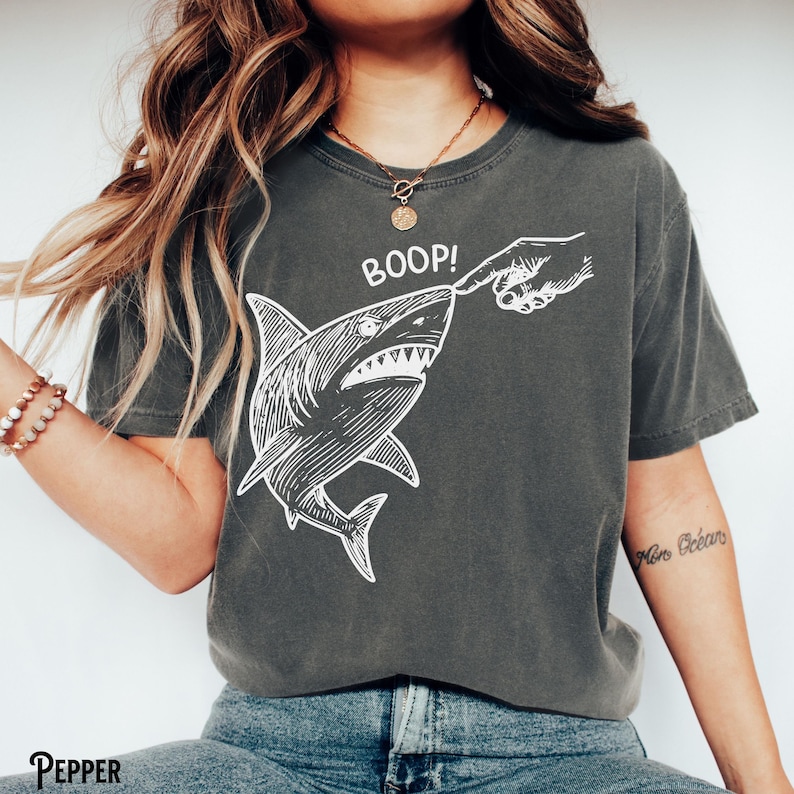 Pu&ograve; includere: T-shirt grigio scuro con un'illustrazione bianca di uno squalo che viene toccato sul naso, con la scritta "BOOP!" sopra. La maglietta ha una vestibilit&agrave; ampia.