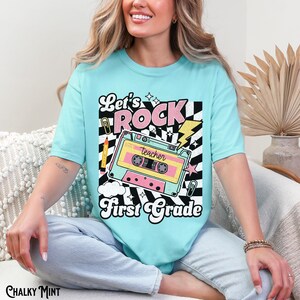May include: A light blue t-shirt with the words "Let's Rock First Grade" in a retro design. The graphic includes a cassette tape, lightning bolt, and a black and white checkered pattern. A pencil and paper clip are also included in the design.