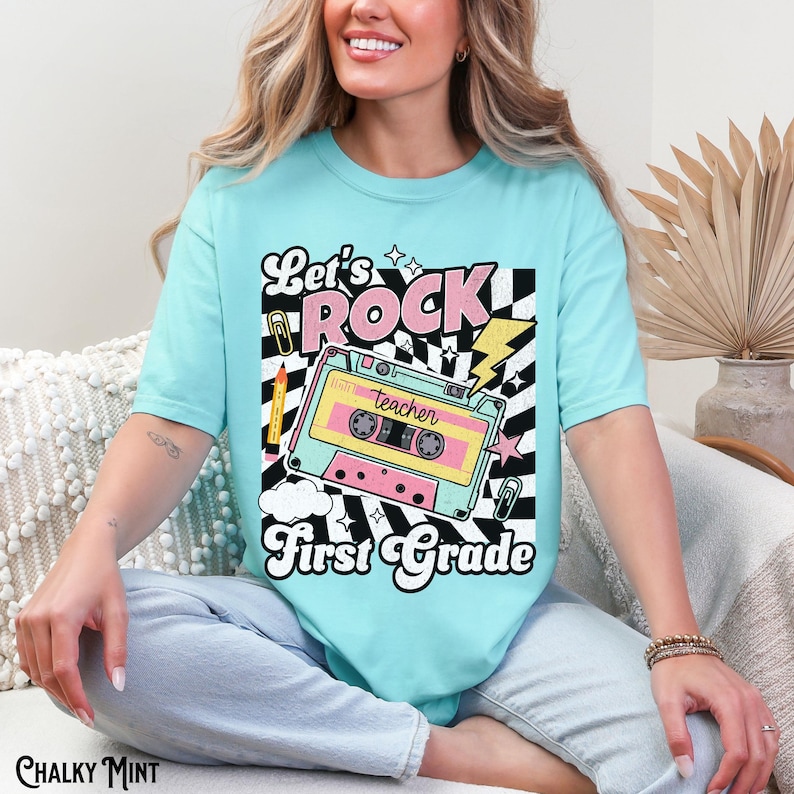 May include: A light blue t-shirt with the words "Let's Rock First Grade" in a retro design. The graphic includes a cassette tape, lightning bolt, and a black and white checkered pattern. A pencil and paper clip are also included in the design.