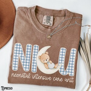 Op de afbeelding: Een bruin T-shirt met de letters "NICU" in een blauw en wit geruit patroon. Een cartoonbeer slaapt op een maan. De woorden "neonatal intensive care unit" staan eronder. Een zilveren ketting ligt over het shirt.