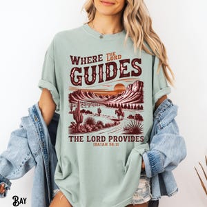 May include: A light green t-shirt with a graphic print of a desert landscape with a sunset and a cowboy riding a horse. The text "Where the Lord Guides, The Lord Provides, Isaiah 58:11" is printed on the shirt.