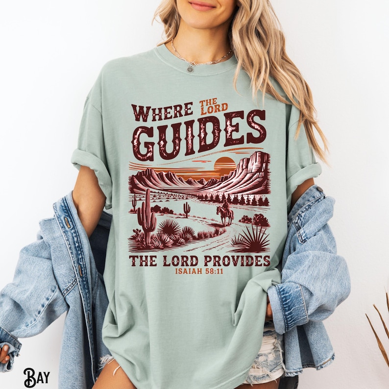 May include: A light green t-shirt with a graphic print of a desert landscape with a sunset and a cowboy riding a horse. The text "Where the Lord Guides, The Lord Provides, Isaiah 58:11" is printed on the shirt.