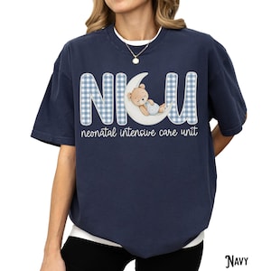 Op de afbeelding: Marineblauwe t-shirt met de letters "NICU" in een blauw ruitpatroon, een slapende teddybeer op een maan en de woorden "neonatal intensive care unit". Het shirt heeft een casual stijl.