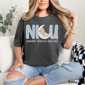 Op de afbeelding: Donkergrijs T-shirt met de letters "NICU" in een blauw ruitpatroon, met een teddybeer op een maan. De woorden "neonatal intensive care unit" staan eronder. Een casual, comfortabele stijl.