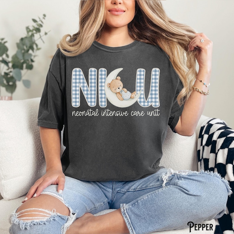 Op de afbeelding: Donkergrijs T-shirt met de letters "NICU" in een blauw ruitpatroon, met een teddybeer op een maan. De woorden "neonatal intensive care unit" staan eronder. Een casual, comfortabele stijl.