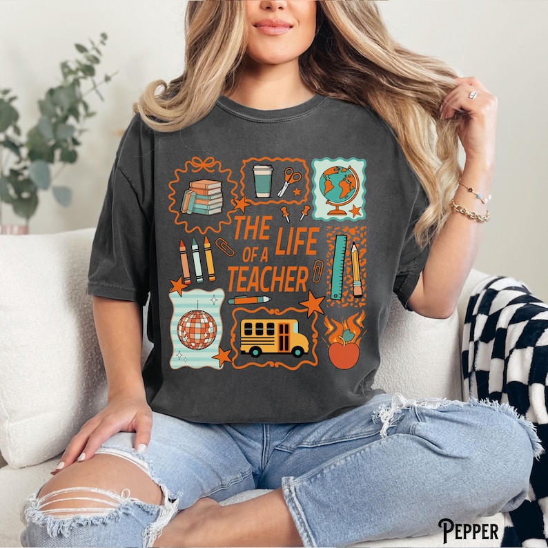 May include: Dark gray t-shirt with a colorful graphic design. The design includes school-related icons such as books, pencils, a globe, and a school bus, along with the text "THE LIFE OF A TEACHER".