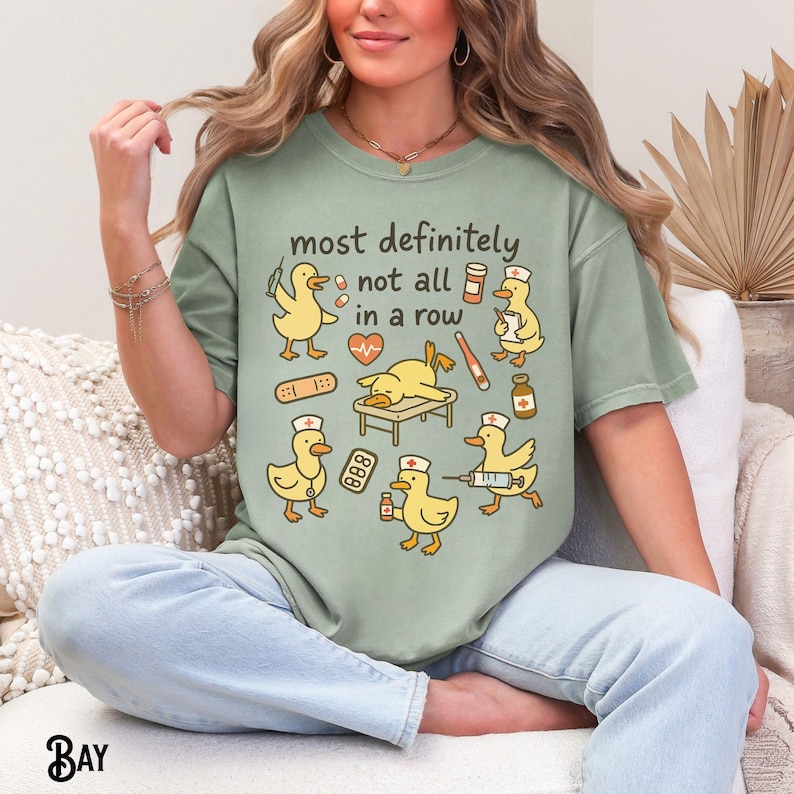 Puede incluir: Camiseta verde salvia con un gr&aacute;fico de patos amarillos de dibujos animados vestidos de enfermeras con suministros m&eacute;dicos. El texto "most definitely not all in a row" est&aacute; encima. La camiseta es de corte holgado.