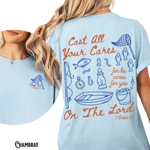 K&ouml;nnte beinhalten: Hellblaues T-Shirt mit einem Angelmotiv. Das Design umfasst ein Fischernetz, Fische, eine Angelrute, ein Boot und den Text "Cast All Your Cares On The Lord 1 Peter 5:7" in Orange und Blau.