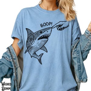 Pu&ograve; includere: T-shirt azzurra con grafica nera di uno squalo che viene toccato sul naso. La parola "BOOP!" &egrave; sopra lo squalo. La maglietta ha maniche corte e una vestibilit&agrave; ampia. Le parole "WASHED DENIM" sono stampate in basso a sinistra.