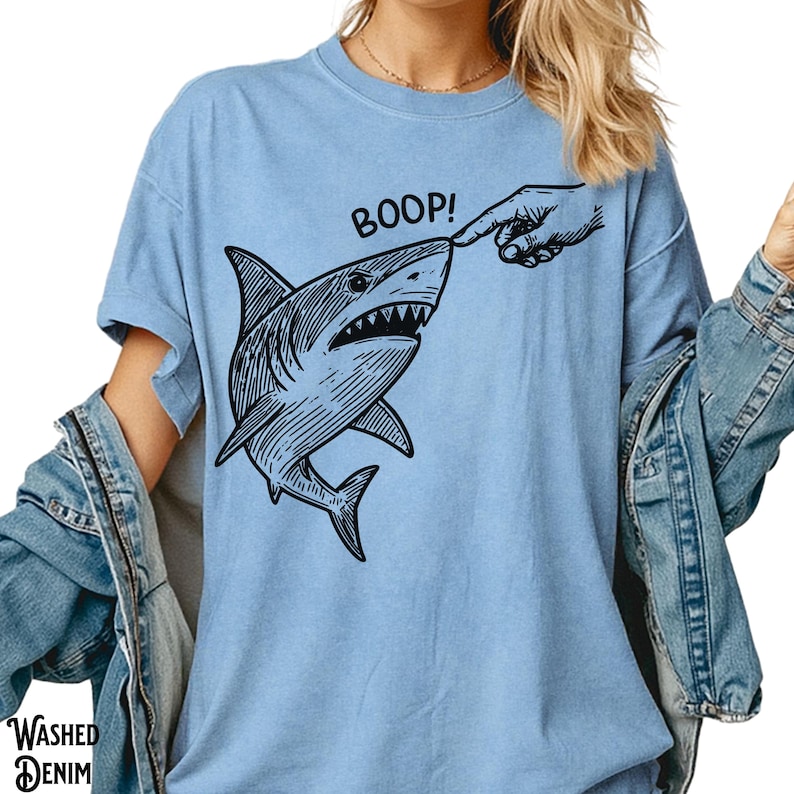 Pu&ograve; includere: T-shirt azzurra con grafica nera di uno squalo che viene toccato sul naso. La parola "BOOP!" &egrave; sopra lo squalo. La maglietta ha maniche corte e una vestibilit&agrave; ampia. Le parole "WASHED DENIM" sono stampate in basso a sinistra.