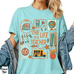 May include: A light blue t-shirt with the text "THE LIFE OF A TEACHER" and various school-themed graphics. The graphics include books, a coffee cup, scissors, a globe, pencils, a ruler, a school bus, and an apple.