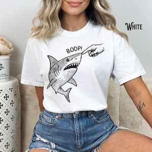 Pu&ograve; includere: T-shirt bianca con un'illustrazione in bianco e nero di uno squalo a cui viene toccato il naso. La parola "BOOP!" &egrave; sopra lo squalo e la parola "White" &egrave; stampata in alto a destra.