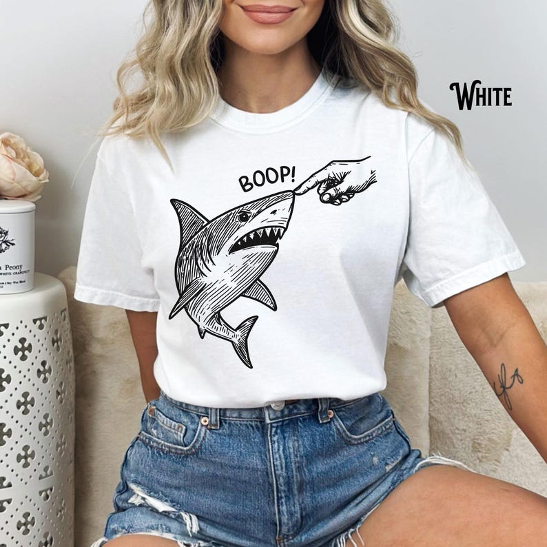 Pu&ograve; includere: T-shirt bianca con un'illustrazione in bianco e nero di uno squalo a cui viene toccato il naso. La parola "BOOP!" &egrave; sopra lo squalo e la parola "White" &egrave; stampata in alto a destra.