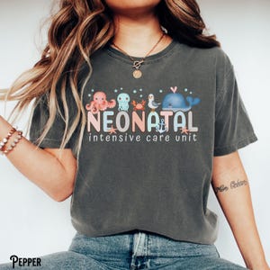 May include: A dark grey t-shirt with a graphic of sea creatures and the text "Neonatal Intensive Care Unit".