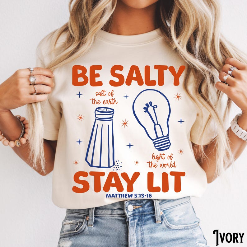 Puede incluir: Camiseta de color marfil con las palabras "BE SALTY STAY LIT" en naranja, junto con ilustraciones azules de un salero y una bombilla. El texto "MATTHEW 5:13-16" est&aacute; impreso debajo. Es una prenda de vestir informal.
