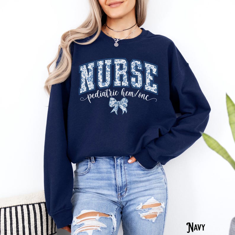 May include: Navy blue sweatshirt with the word "NURSE" in a floral design. Below the word "NURSE" is the text "pediatric hem/onc" and a floral bow. The word "NAVY" is in the bottom right corner.
