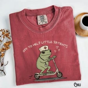 May include: A maroon Comfort Colors t-shirt featuring a cartoon frog wearing a nurse's hat and riding a scooter. The text "OFF TO HELP LITTLE PATIENTS" is above the frog. A small tag reads "S P CH".