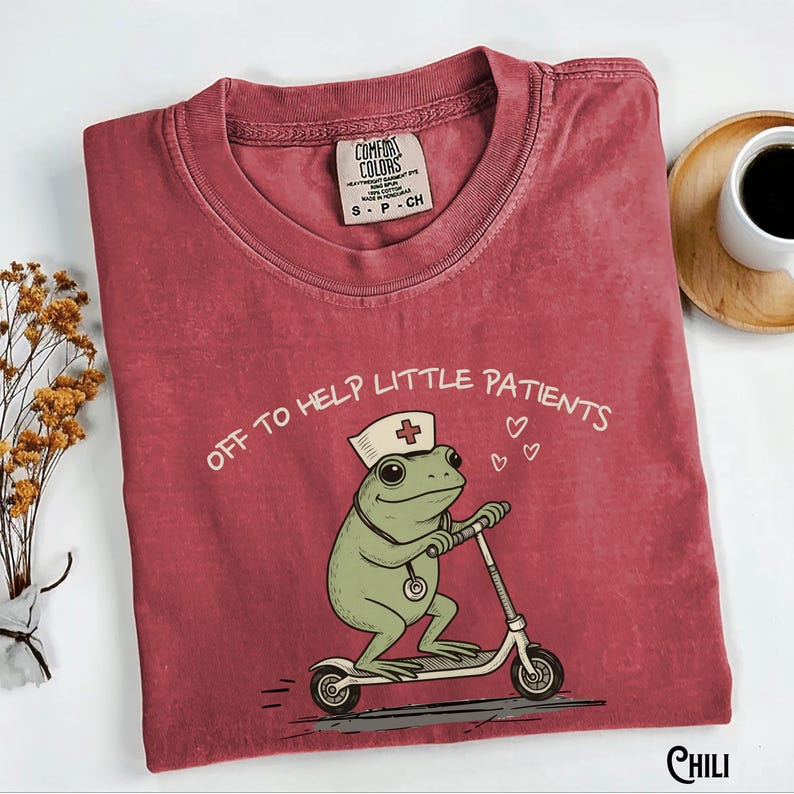 May include: A maroon Comfort Colors t-shirt featuring a cartoon frog wearing a nurse's hat and riding a scooter. The text "OFF TO HELP LITTLE PATIENTS" is above the frog. A small tag reads "S P CH".
