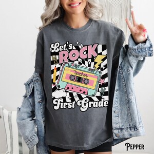 May include: Dark gray t-shirt with a retro design featuring the words "Let's ROCK First Grade." The graphic includes a cassette tape, lightning bolt, and a black and white checkered pattern. A pencil and paperclip are also included in the design.