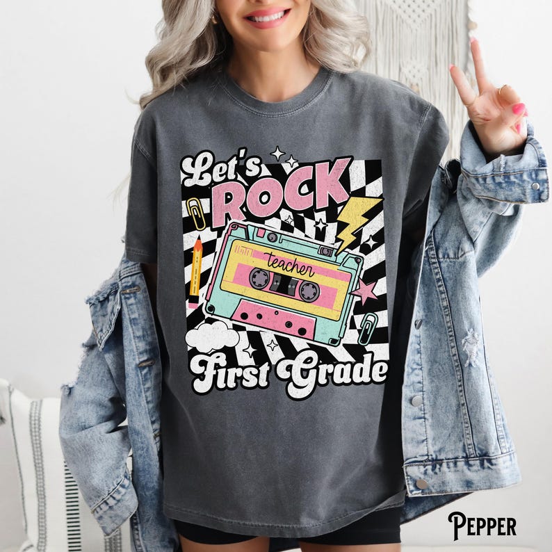 May include: Dark gray t-shirt with a retro design featuring the words "Let's ROCK First Grade." The graphic includes a cassette tape, lightning bolt, and a black and white checkered pattern. A pencil and paperclip are also included in the design.