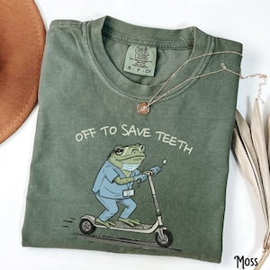 May include: A moss green t-shirt featuring the text "OFF TO SAVE TEETH". The graphic shows a frog in scrubs, a face mask, and glasses, riding a scooter. A gold necklace is draped across the shirt. The shirt is size S-P-CH.
