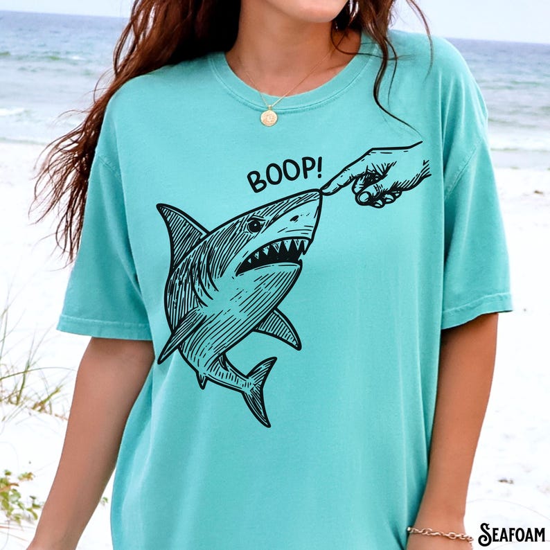 Pu&ograve; includere: T-shirt verde acqua con grafica nera di uno squalo a cui viene toccato il naso con un dito, con la scritta "BOOP!" sopra. La maglietta &egrave; a tinta unita e ha maniche corte.