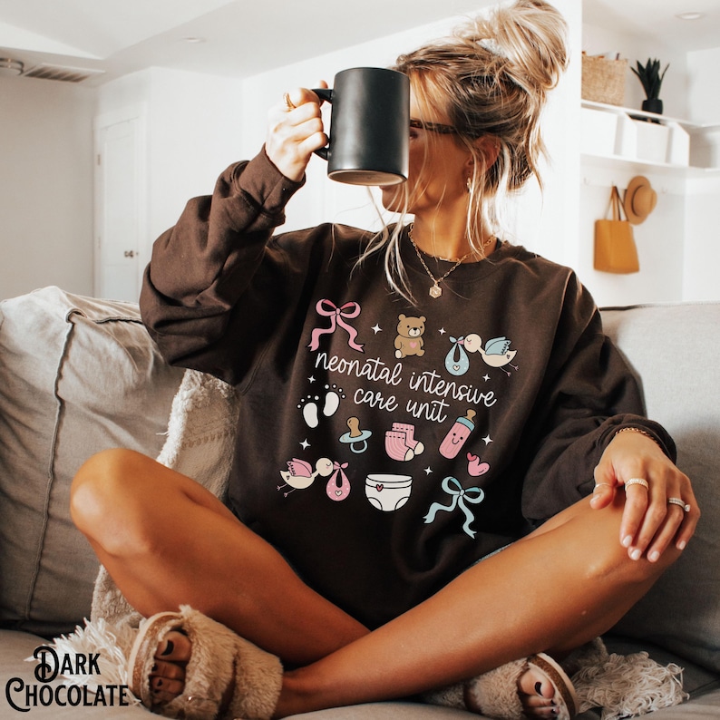 Op de afbeelding: Een bruin sweatshirt met de tekst "neonatal intensive care unit" en verschillende baby-iconen in roze, blauw en wit.