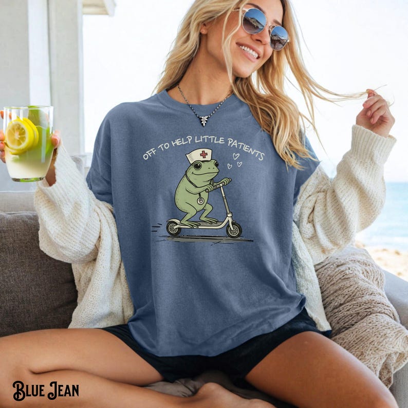 May include: Blue jean colored t-shirt with a cartoon frog wearing a nurse's hat and riding a scooter. The text "OFF TO HELP LITTLE PATIENTS" is printed above the frog. The shirt is a relaxed fit.