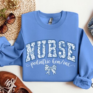 May include: Carolina blue sweatshirt with the word "NURSE" in a floral design. Below the word "NURSE" is the text "pediatric hem/onc" and a bow graphic. The sweatshirt is paired with brown shoes and sunglasses.