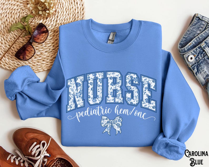 May include: Carolina blue sweatshirt with the word "NURSE" in a floral design. Below the word "NURSE" is the text "pediatric hem/onc" and a bow graphic. The sweatshirt is paired with brown shoes and sunglasses.