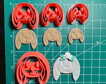 May include: A set of five cookie cutters in the shape of bunnies. The cutters are made of red, brown, and gray plastic. The cutters are arranged on a green cutting mat with a ruler in inches.