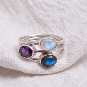 May include: A stack of three silver rings, each featuring an oval-shaped gemstone. The top ring has a translucent white moonstone, the middle ring holds a deep purple amethyst, and the bottom ring displays a dark blue labradorite gemstone.